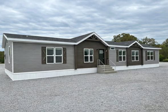 A modern, single-story modular home with brown siding, multiple white-framed windows, and a central front entrance with steps, set against a cloudy sky.