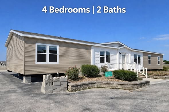 Single-story beige modular home with white trim, four windows, and a front porch. Nearby are bushes and a short brick wall. Text reads "4 Bedrooms | 2 Baths."