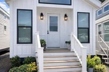 A small, white tiny house with a gabled roof and porch. Surrounded by greenery, it has rectangular windows and hanging lights. Price details: $8000 full, $500 down, financing available.