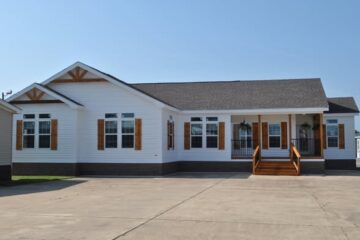 A modern white mobile home with wooden shutters and a gabled roof. It features a front porch with steps and a smooth concrete driveway under a clear sky.