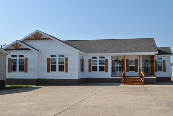 A modern white mobile home with wooden shutters and a gabled roof. It features a front porch with steps and a smooth concrete driveway under a clear sky.