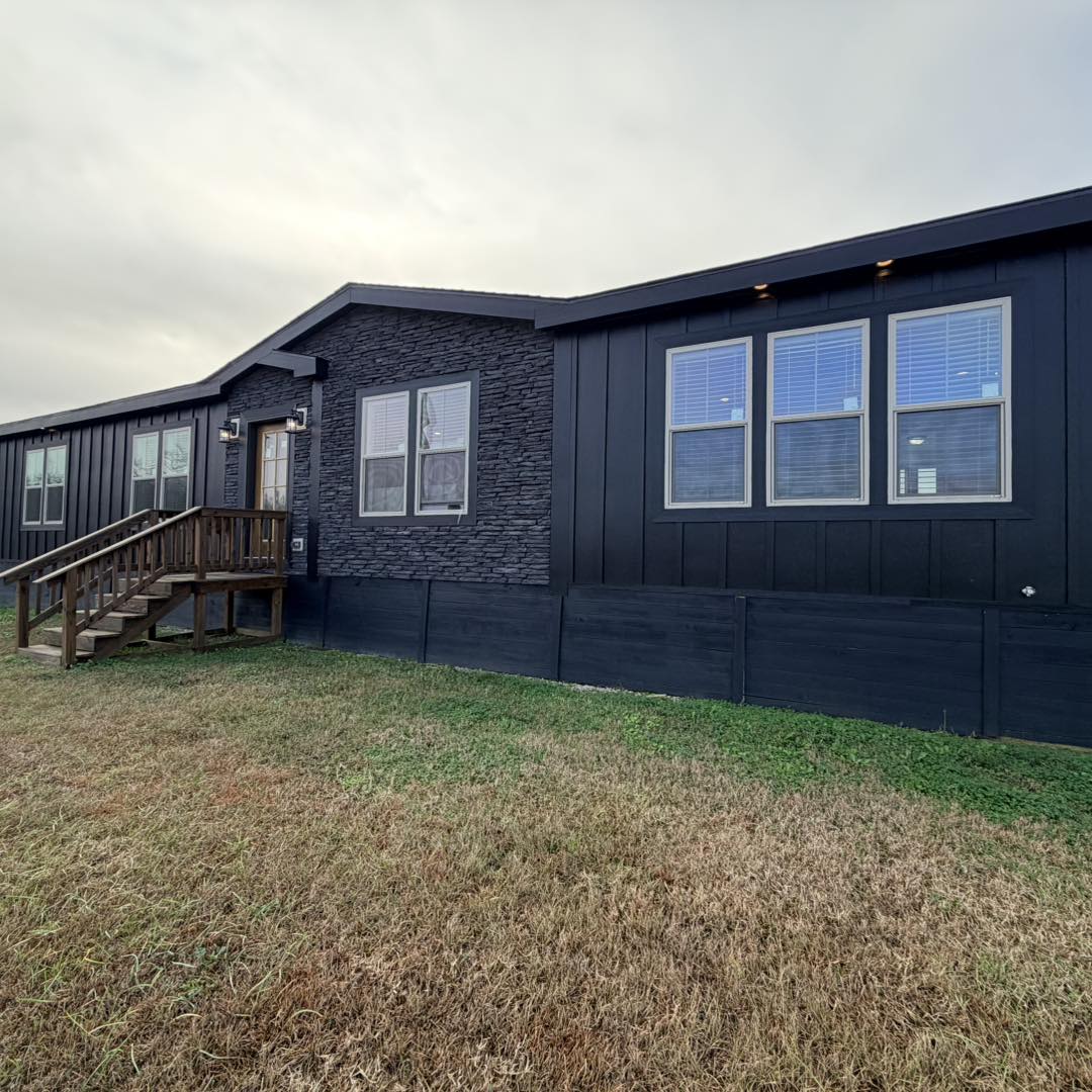 Modern black modular home with a wooden porch, gray stone accents, and multiple windows. It sits on a grassy lawn under a cloudy sky.