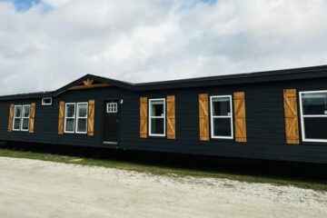 Long, single-story, dark gray modular home with wooden shutters and white-trimmed windows. It's set against a cloudy sky and gravel ground.