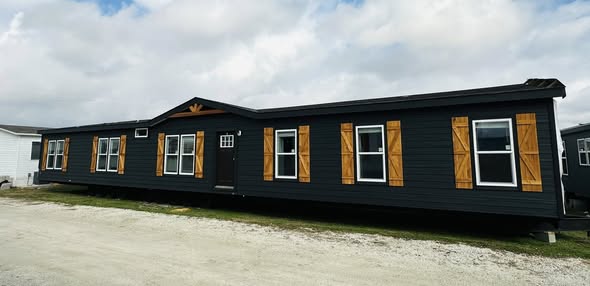 Long, single-story, dark gray modular home with wooden shutters and white-trimmed windows. It's set against a cloudy sky and gravel ground.
