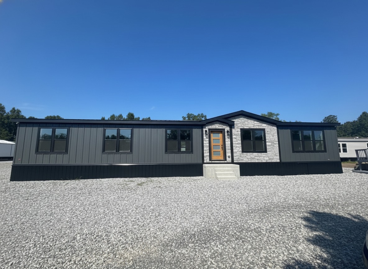 Modern gray modular home with a stone facade, large windows, and a wooden door, situated on a gravel lot under a clear blue sky.