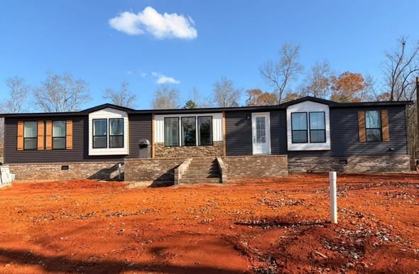 A single-story modular home with a dark facade and large windows sits on red clay soil under a clear blue sky, evoking a sense of tranquility.