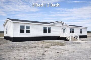 Single-story white manufactured home on a gravel plot, featuring large windows and a small porch. Text reads "3 Bed | 2 Bath." Sky is partly cloudy.