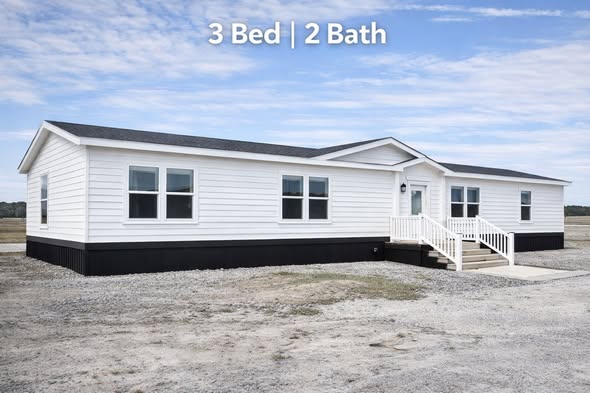 Single-story white manufactured home on a gravel plot, featuring large windows and a small porch. Text reads "3 Bed | 2 Bath." Sky is partly cloudy.