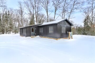 A modern, dark gray cabin sits amidst a snowy landscape surrounded by bare trees, evoking a serene, wintery feel. Bright sky contrasts with the snow.