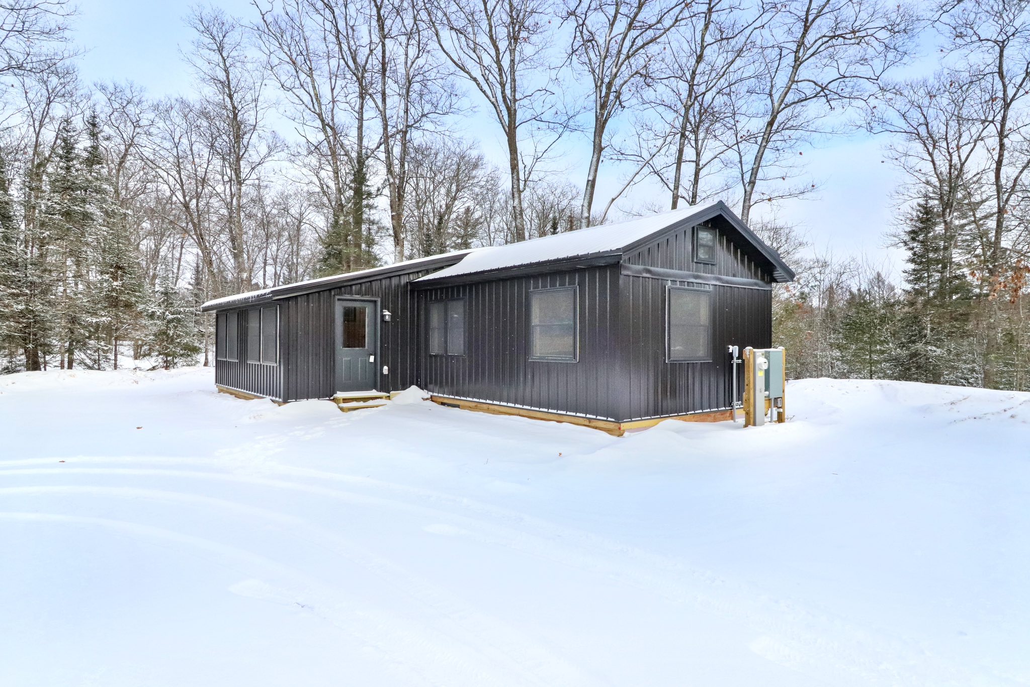 A modern, dark gray cabin sits amidst a snowy landscape surrounded by bare trees, evoking a serene, wintery feel. Bright sky contrasts with the snow.