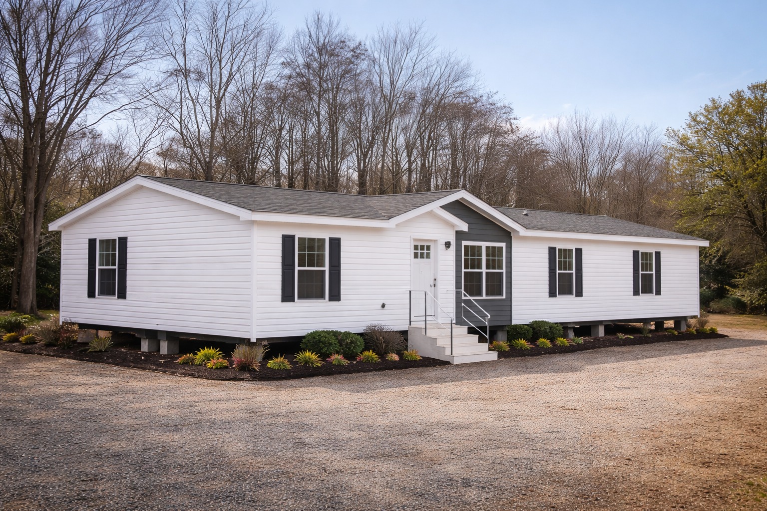 A white prefabricated house with black shutters sits elevated on a gravel lot. Sparse landscaping with shrubs surrounds the home. Trees line the background.
