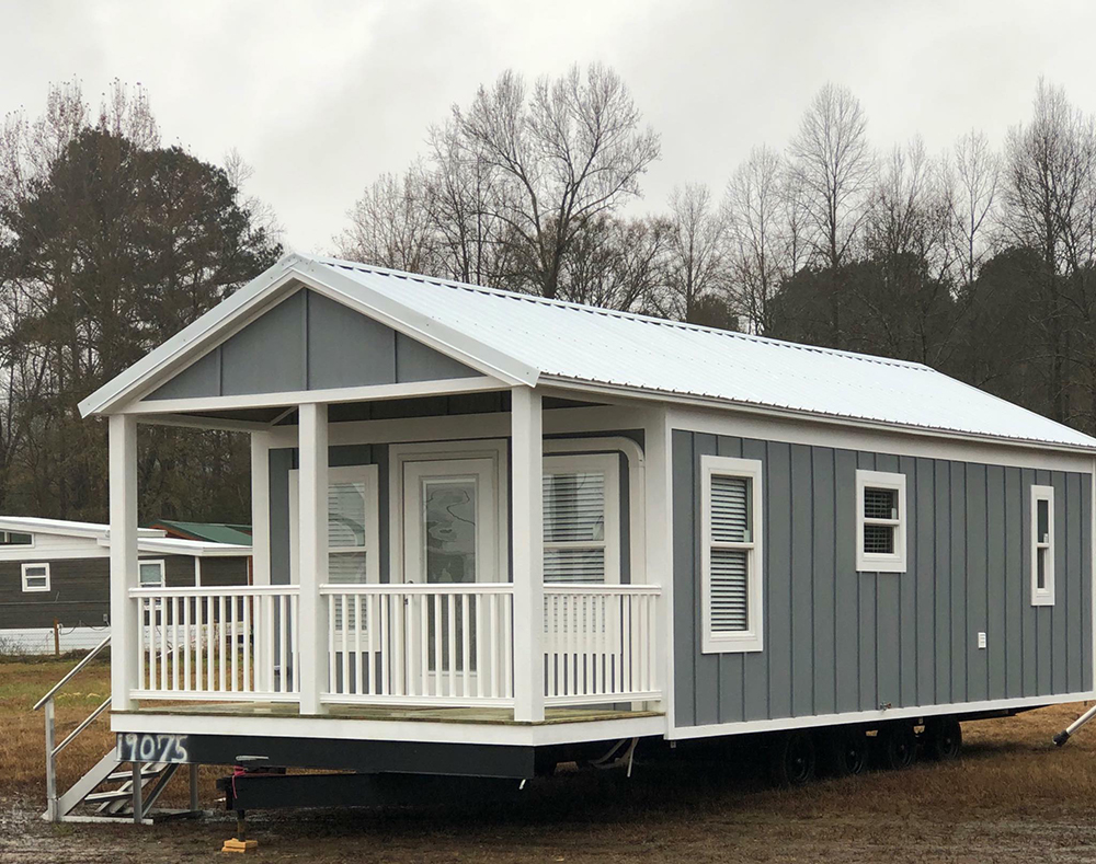 A small, gray and white tiny house on wheels with a front porch and steps, set in a grassy area with leafless trees in the background, under an overcast sky.