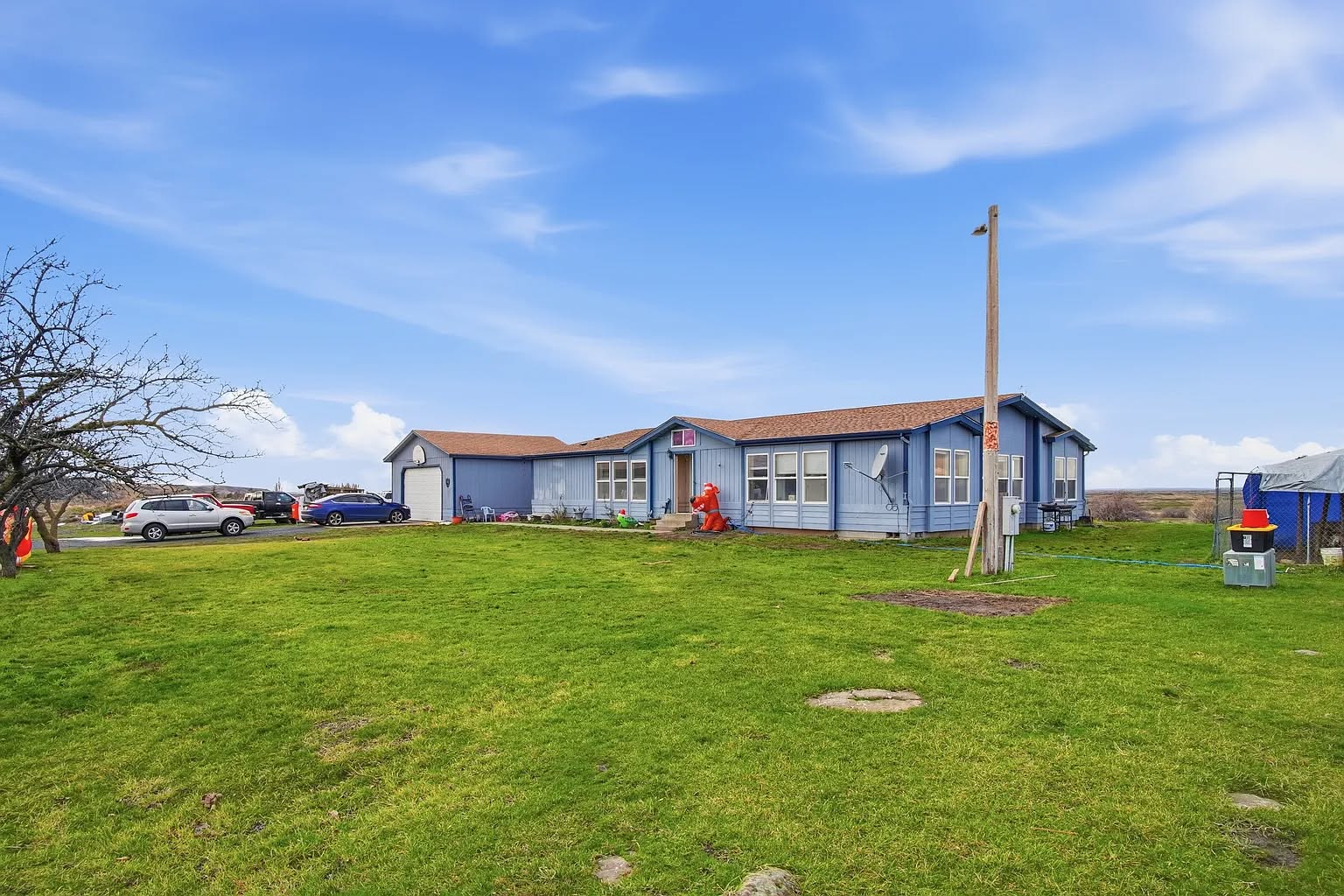 A blue house with a brown roof sits on a lush green lawn under a bright blue sky. Cars are parked nearby, with a solitary leafless tree on the left.