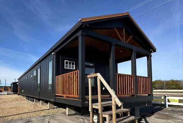 A modern black tiny house with a front porch, wooden railings, and a peaked roof. The house sits on a gravel lot under a clear blue sky.