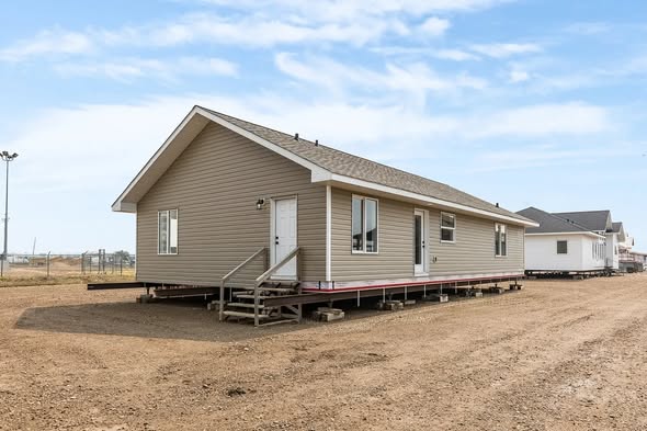 A beige modular home is situated on a dirt lot under a clear blue sky. The house has steps leading to the door, with windows along the sides.