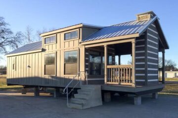 Small wooden cabin with a metal roof and raised porch, set on concrete blocks. The structure is bathed in warm sunlight against a clear blue sky.