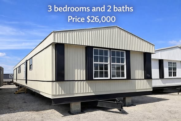 Manufactured home with beige siding, white-framed windows, and black trim sits on gravel under a blue sky. Text reads: "3 bedrooms and 2 baths, Price $26,000."