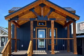 Compact tiny house with blue siding and natural wood accents, front porch, and stairs. Text reads "1 bed 1 bath 399 sqft" against a clear sky.