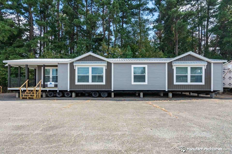 Gray manufactured home with a green roof on a gravel lot, surrounded by tall pine trees. Three large windows and small porch with steps at one end.