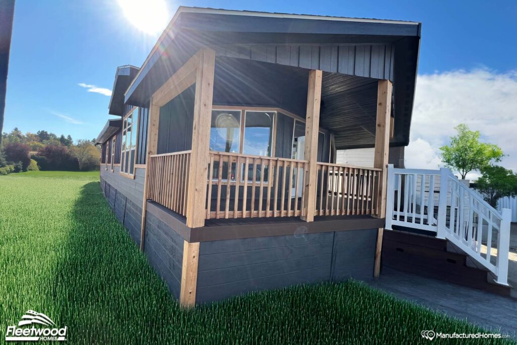 A modern manufactured home with a wooden porch and railing stands on a lush green lawn under a bright blue sky. The sun creates a warm, inviting glow.
