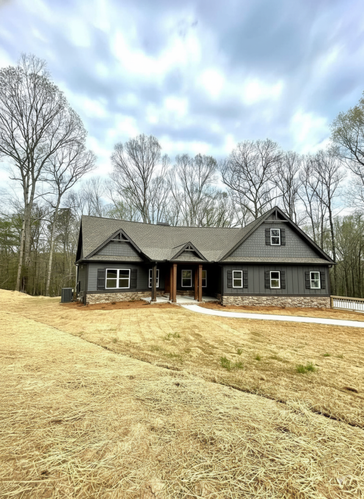 A gray single-story house with a gabled roof sits on a straw-covered lot, surrounded by leafless trees under a partly cloudy sky. A serene, rural setting.