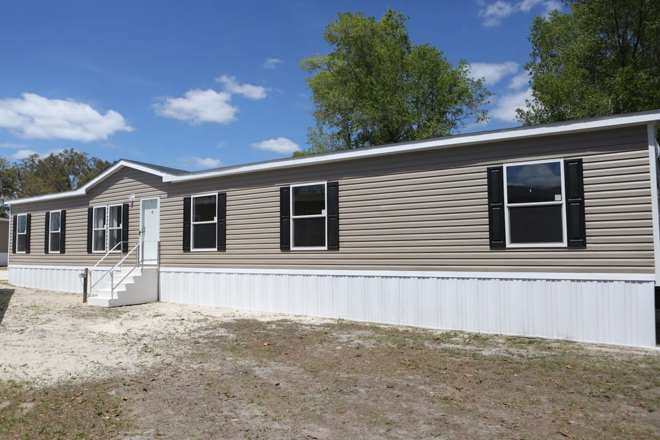 Single-story mobile home with beige siding and black shutters. It sits on sandy ground under a blue sky with scattered clouds and green trees.