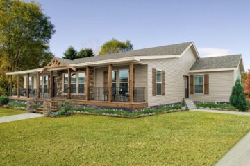 A single-story house with beige siding, a large wooden porch, and black railings. Surrounded by a green lawn and trees under a clear sky.