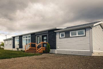 Modern manufactured home under a cloudy sky, featuring gray siding, black accents, and wooden steps leading to the entrance, set on a gravel driveway.