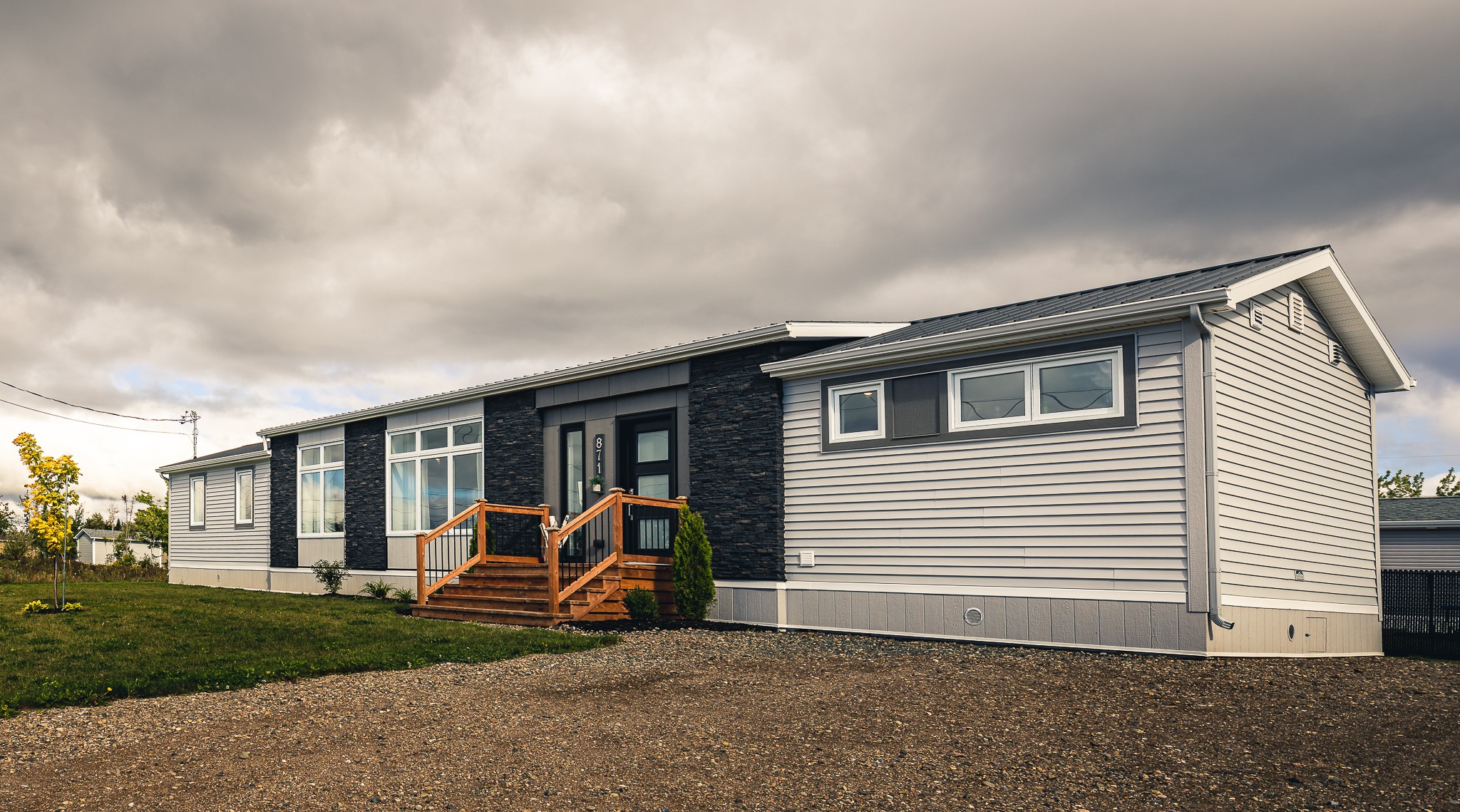 Modern manufactured home under a cloudy sky, featuring gray siding, black accents, and wooden steps leading to the entrance, set on a gravel driveway.