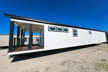 Modern tiny home with white exterior and several small windows, elevated on a support. It features a covered porch with black railings under a clear blue sky.