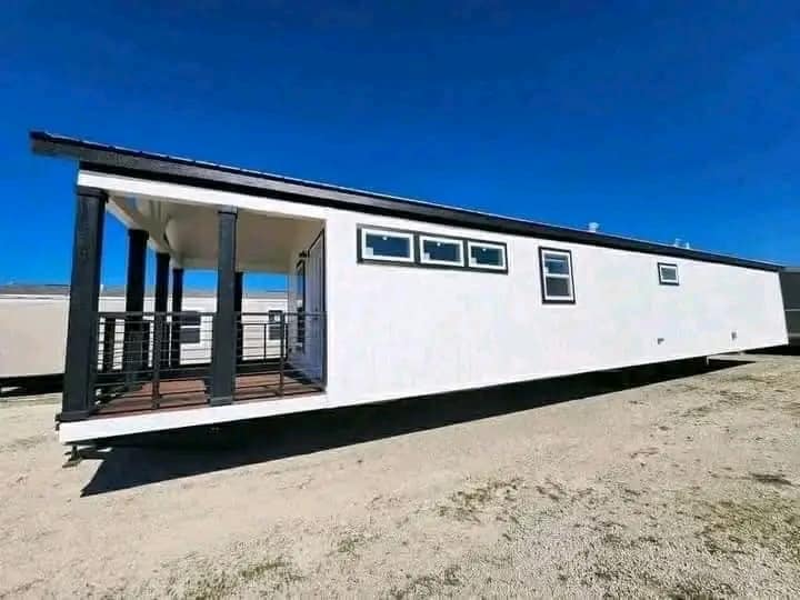 Modern tiny home with white exterior and several small windows, elevated on a support. It features a covered porch with black railings under a clear blue sky.