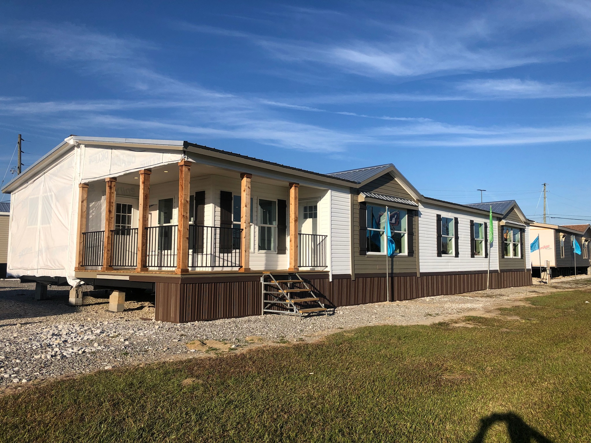 Large manufactured home on display, featuring a wide porch with wooden columns and neat steps. It sits on a gravel lot under a bright blue sky.