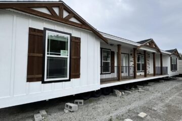 Modular home with white siding and wooden trim, elevated on concrete blocks, under a cloudy sky. The house features large windows and a small porch.