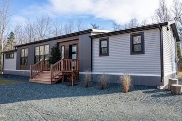 Modern manufactured home with gray siding and dark trim, set on gravel with wooden steps leading to the entrance. Bare trees stand in the background.