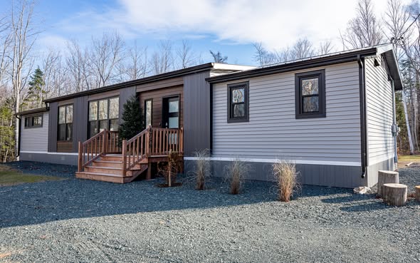 Modern manufactured home with gray siding and dark trim, set on gravel with wooden steps leading to the entrance. Bare trees stand in the background.