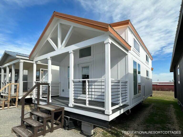 A modern tiny house with a white facade and brown roof under a clear blue sky. It features a small porch with railings and steps leading up.