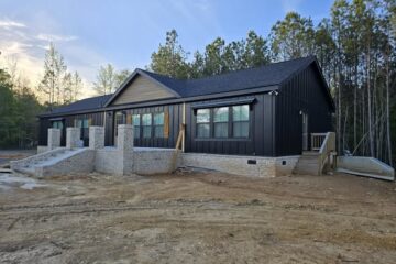 Modern black house with large windows, under construction. Earth-toned brick foundation, surrounded by dirt and pine trees, under a blue sky.