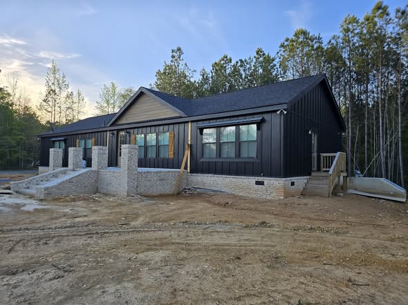 Modern black house with large windows, under construction. Earth-toned brick foundation, surrounded by dirt and pine trees, under a blue sky.