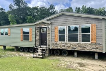 A small manufactured home with gray siding, stone accents, and wooden shutters. It has multiple windows, a black door with steps, and sits on grass.