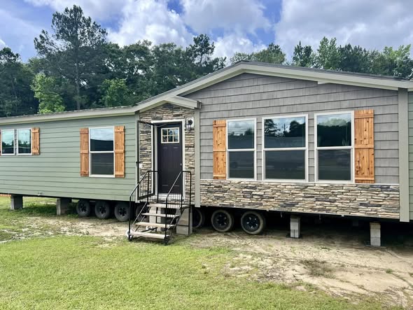 A small manufactured home with gray siding, stone accents, and wooden shutters. It has multiple windows, a black door with steps, and sits on grass.