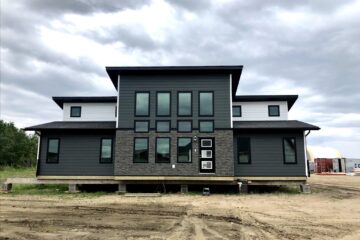Modern, two-story house with dark gray siding and large windows, set on a dirt lot under a cloudy sky, conveying a contemporary and sturdy feel.