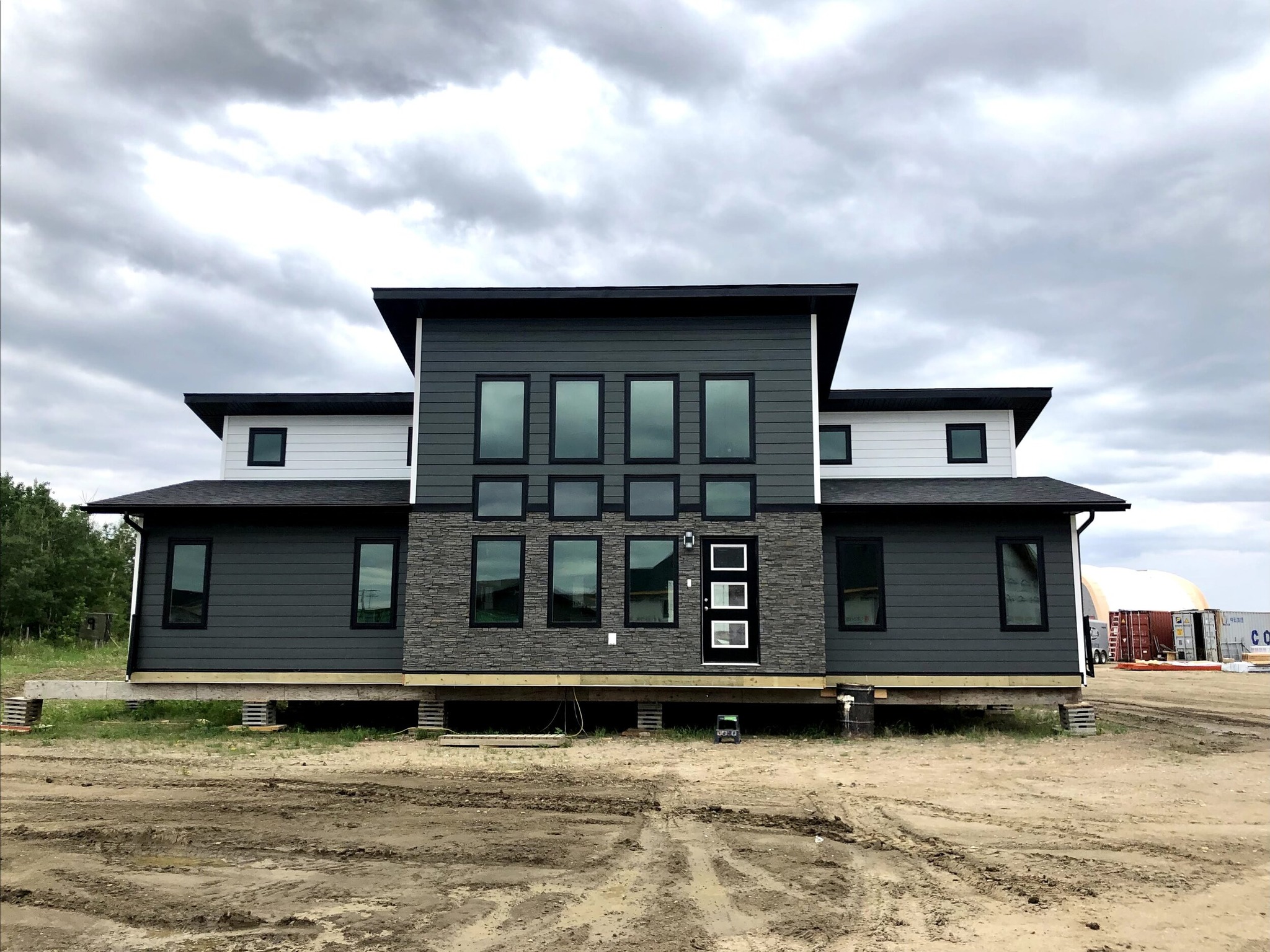 Modern, two-story house with dark gray siding and large windows, set on a dirt lot under a cloudy sky, conveying a contemporary and sturdy feel.