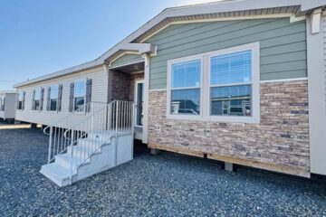 A modern manufactured home with green siding and brick accents. Features include a set of steps leading to the entrance, and multiple windows.