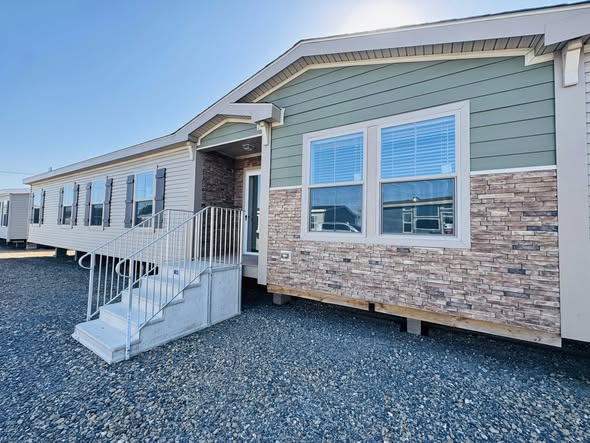 A modern manufactured home with green siding and brick accents. Features include a set of steps leading to the entrance, and multiple windows.