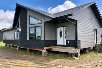 A modern, prefabricated house with gray siding and dark stone accents sits elevated on blocks in a muddy field under a bright blue sky with clouds.
