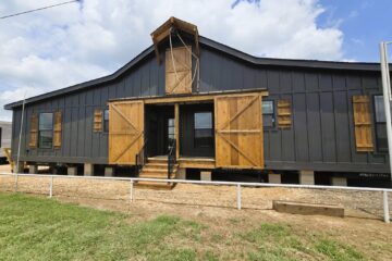 A modern, barn-style building with dark gray siding and large wooden shutters. It features a porch and sits on a raised foundation under a bright blue sky.