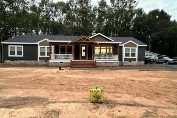 A single-story house with a porch and peaked roof is under construction, set against a backdrop of tall trees. The surrounding area is bare soil.