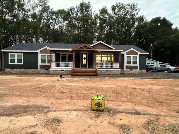 A single-story house with a porch and peaked roof is under construction, set against a backdrop of tall trees. The surrounding area is bare soil.