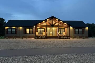 Single-story house at dusk, softly lit with warm lights. It has a gabled roof, large porch, and stone pathway, creating a welcoming, serene atmosphere.