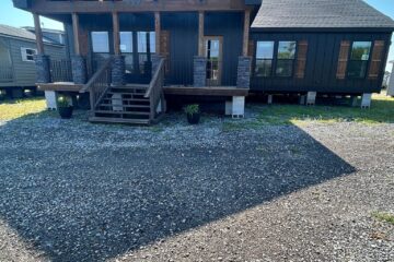 A modern cabin with a wooden porch and exposed beams under a clear blue sky. Gravel pathway leads to steps with potted plants. Calm and rustic.